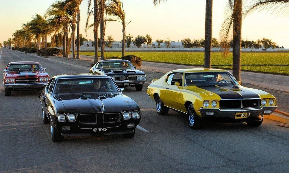 Three American muscle cars, including a black Pontiac GTO and a yellow muscle car, parked on a palm-lined road