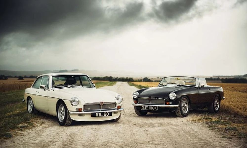 Two classic sports cars, one cream and one black, parked on a dirt road under a cloudy sky
