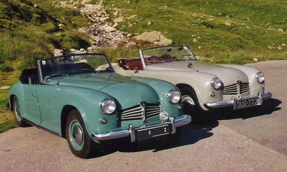 Two vintage convertibles, one green and one cream, parked on a country road