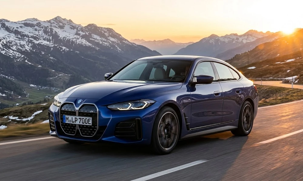 Blue BMW i4 driving on a mountain road with snowy peaks and a rising sun behind it