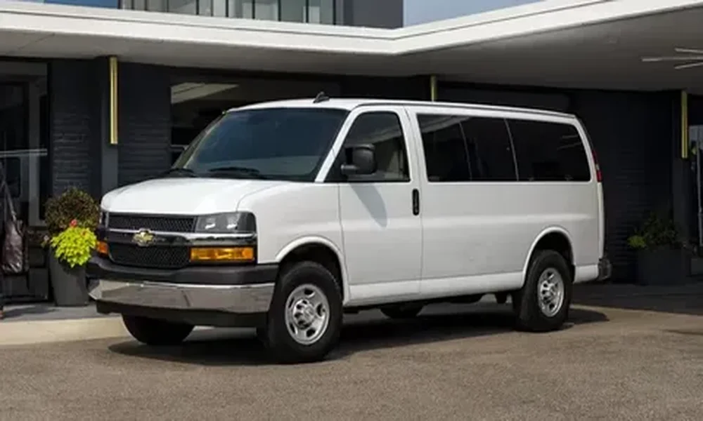 White Chevrolet Express passenger van with tinted windows, parked in front of a modern building entrance.