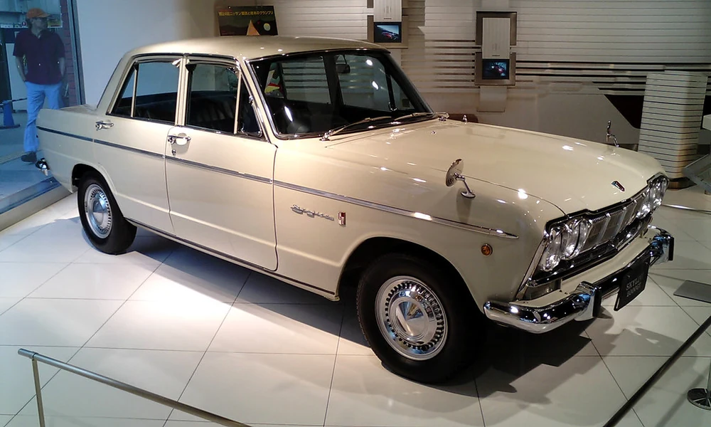Cream-colored early Nissan Skyline four-door sedan with chrome trim and a vintage front grille, displayed indoors.