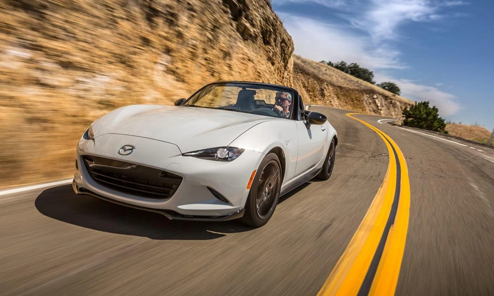 White Mazda MX-5 Miata with the roof down cornering on a road beside a rocky hillside