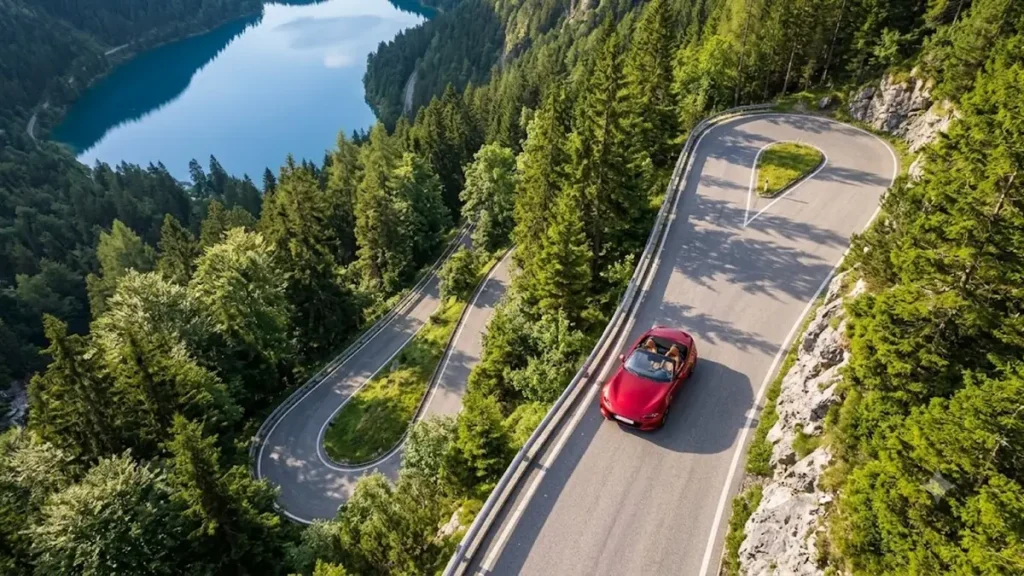 Aerial view of a red convertible with the roof down driving on a winding mountain road beside a blue lake
