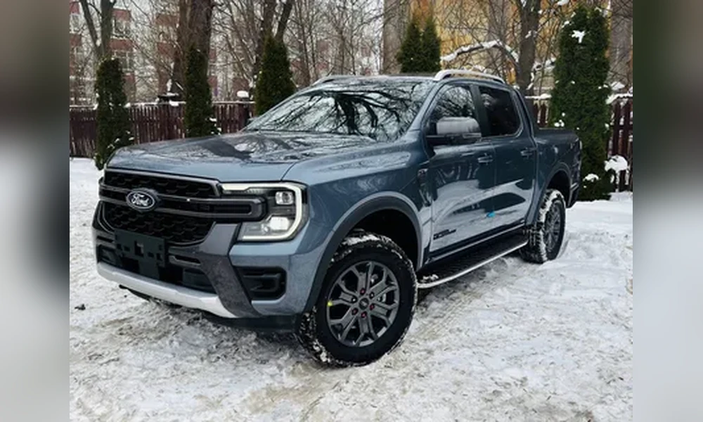 Blue-gray Ford Ranger double cab parked on snow with bare trees and a fence behind it