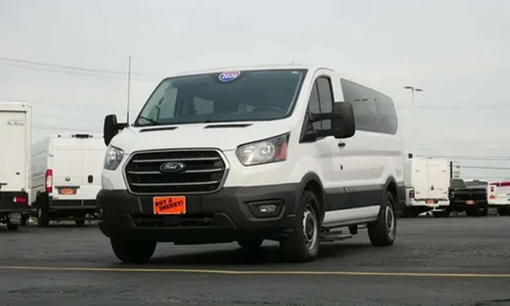 White Ford Transit passenger van with tinted rear windows, parked in a lot with other vans in the background.
