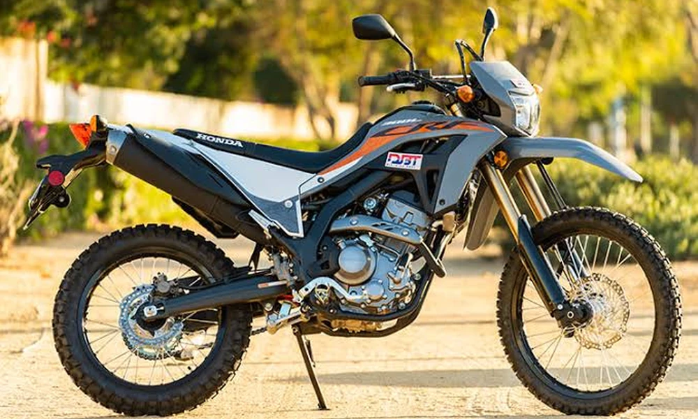 Gray and orange Honda CRF300L dual-sport motorcycle with knobby tires, side view on a sandy dirt road with trees in the background