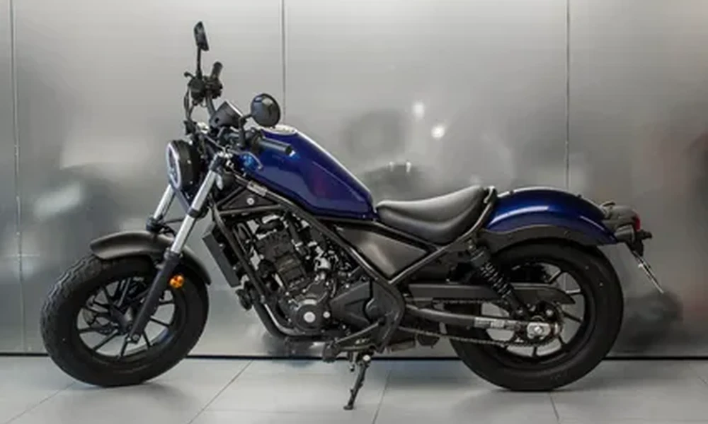 Blue Honda Rebel 300 cruiser motorcycle with black seat and spoked wheels, side view in front of a brushed metal wall