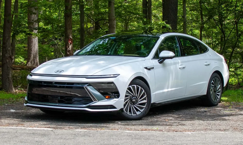 White Hyundai Sonata Hybrid sedan parked on gravel in front of a forest