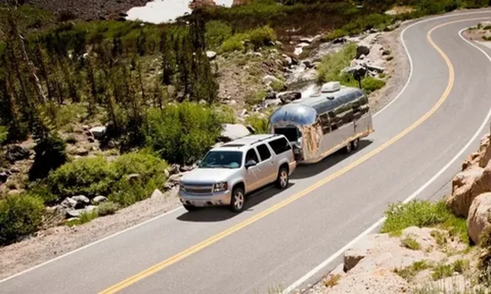 White full-size SUV pulling a silver travel trailer around a curve on a paved mountain road, with trees and rocky slopes alongside.