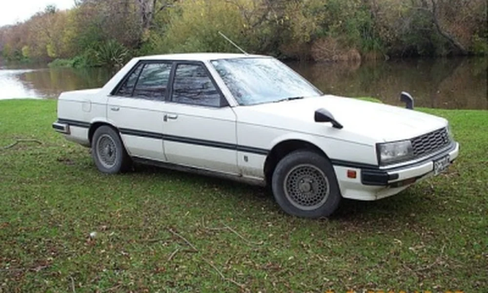 White Nissan Skyline R30 four-door sedan parked on grass beside a river, featuring rectangular headlights and classic 1980s styling.