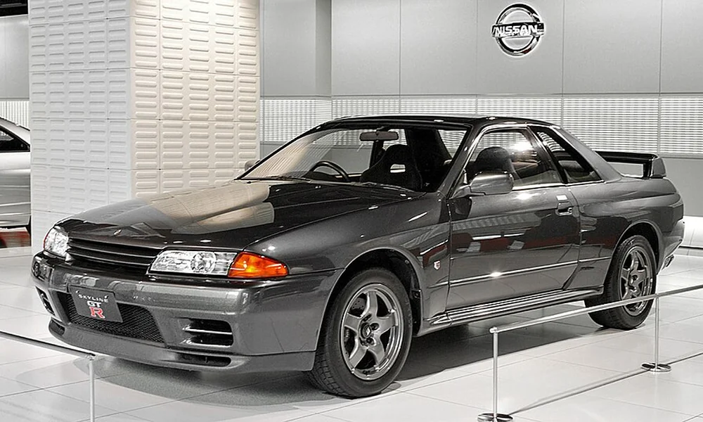 Dark grey Nissan Skyline R32 GT-R coupe with a rear spoiler, alloy wheels, and distinctive front headlights in a showroom.