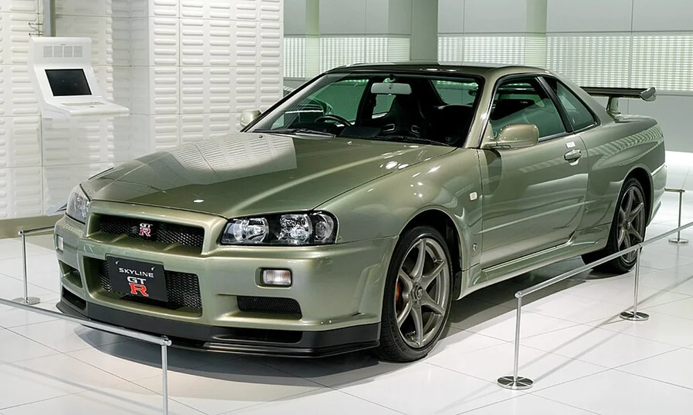 Olive-green Nissan Skyline R34 GT-R coupe with a rear spoiler, alloy wheels, and distinctive front grille, displayed indoors.