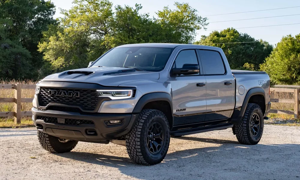 Gray Ram 1500 with black wheels parked on gravel beside a wooden fence