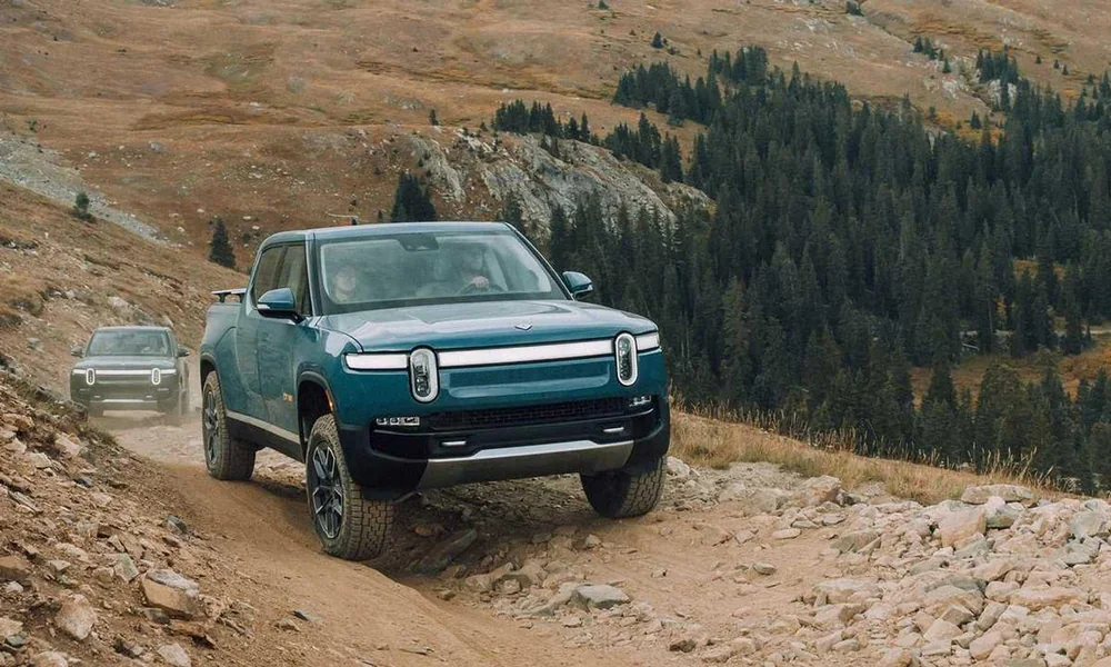 Rivian R1T electric pickup truck driving along a gravel trail in a forested landscape.