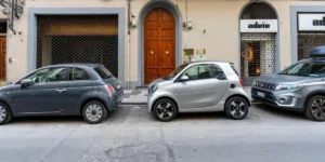 Three smart cars parked in a row at the curb in front of a beige building with an arched wooden door, with the smallest silver one in the middle