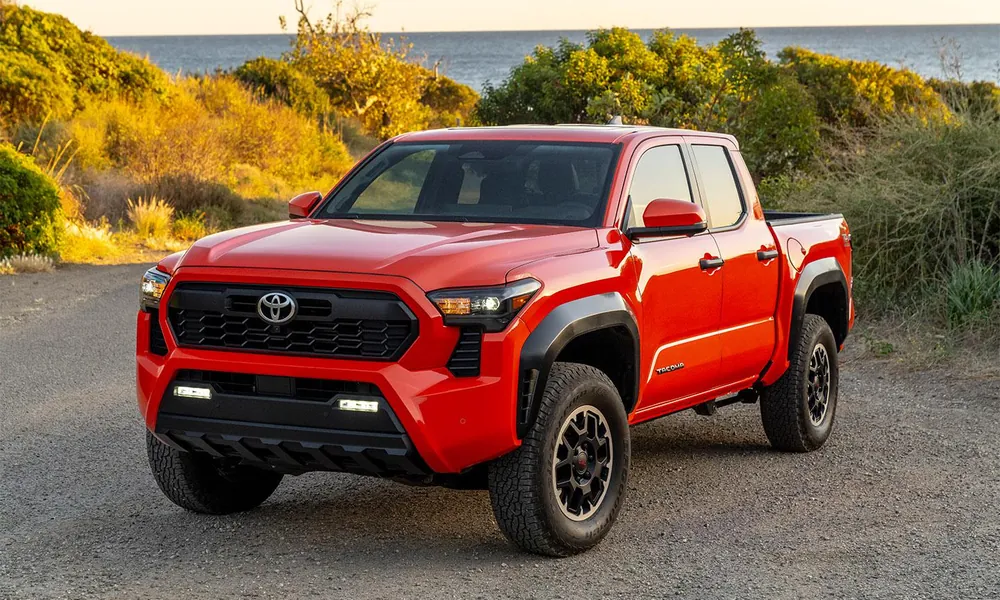 Red Toyota Tacoma midsize pickup parked on gravel with coastal brush behind it